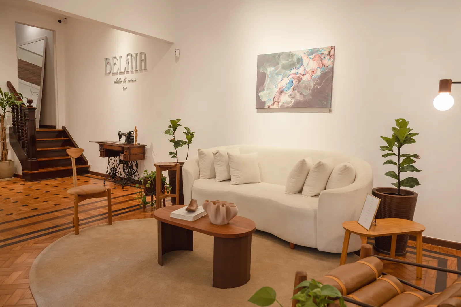 Sala da Casa Belina, com sofá branco, piso de taco antigo e o letreiro do ateliê na parede