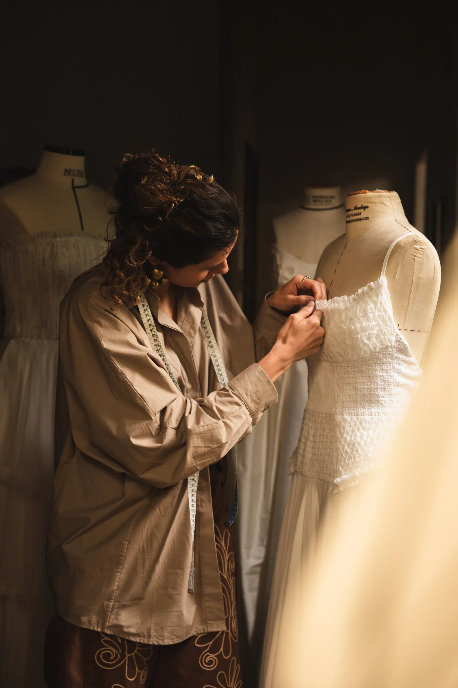 Gabriella Moura, a Gabi, com fita métrica no pescoço, ajustando o corpete de um vestido branco no manequim do ateliê