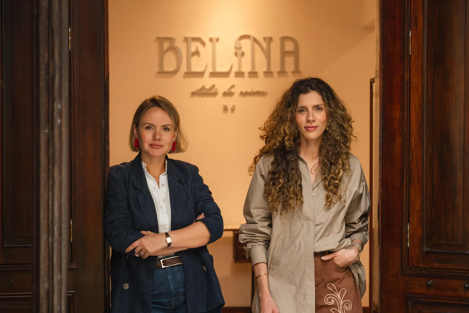 Leidi Taveira, à esquerda, e Gabriella Moura, à direita, sócias da Belina, de pé entre as portas de madeira do salão, com o letreiro do ateliê na parede ao fundo